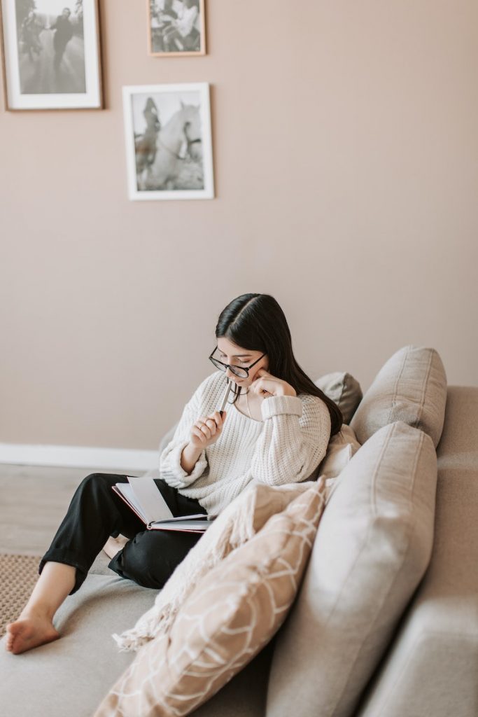 Concentrated businesswoman sitting barefoot with legs crossed on comfortable sofa in modern living room and planning business project during remote work while touching cheek and lips with pen